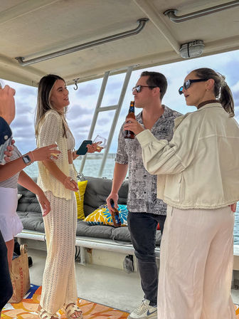Friends chatting and sipping drinks on a covered boat deck in coastal waters — casual summer outfits, colorful cushions and ladder visible against a cloudy sky backdrop