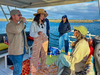 Group of friends socializing on a covered boat at a coastal marina, wearing sun hats and casual clothes, colorful cushions and rugs, holding drinks with a rocky breakwater and calm ocean in the background.
