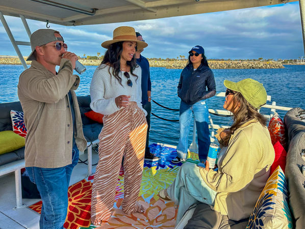 Group of friends socializing on a covered boat at a coastal marina, wearing sun hats and casual clothes, colorful cushions and rugs, holding drinks with a rocky breakwater and calm ocean in the background.