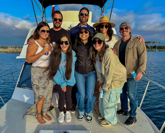 Cheerful group of eight friends posing on the bow of a sunlit boat, wearing sunglasses and casual summer outfits, holding canned drinks with a calm marina and palm trees on the horizon under a blue sky.