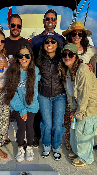 Cheerful group of eight friends posing on the bow of a sunlit boat, wearing sunglasses and casual summer outfits, holding canned drinks with a calm marina and palm trees on the horizon under a blue sky.