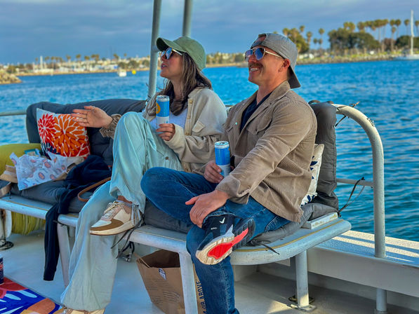 A man and woman in caps and sunglasses relax with canned drinks on a boat bench, smiling as they overlook blue harbor waters and a palm-lined coastal marina.
