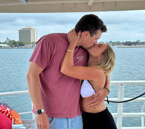 Romantic couple kissing on a boat deck, woman in a white bikini top and man in a maroon T-shirt embracing with calm harbor water and a coastal city skyline in the background.