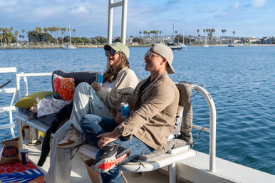 Two people in caps and sunglasses relax on a boat bench holding canned drinks, smiling toward a palm-lined shoreline with sailboats in a sunny coastal marina.