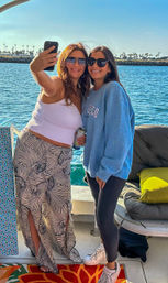 Two smiling friends taking a selfie on a sunny boat near a palm-tree lined harbor, blue water and clear sky in the background.