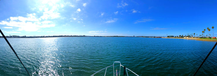 Panoramic view from a boat bow across a sunlit coastal bay with sparkling blue water, sandy shoreline dotted with palm trees and a clear blue sky.