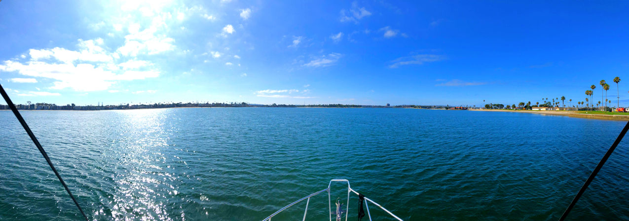 Panoramic view from a boat bow across a sunlit coastal bay with sparkling blue water, sandy shoreline dotted with palm trees and a clear blue sky.