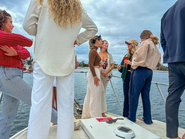 Group of people socializing on a boat deck near a coastal marina under a cloudy sky, wearing casual outfits and holding drinks.