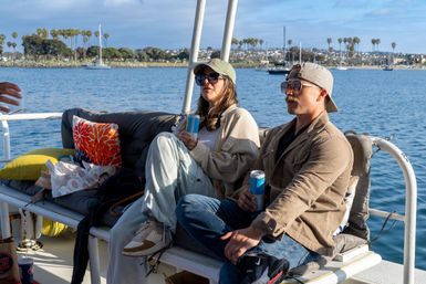 Two people in caps and sunglasses relax on a cushioned bench of a small boat, sipping canned drinks with sailboats and palm trees along a sunny coastal harbor in the background.
