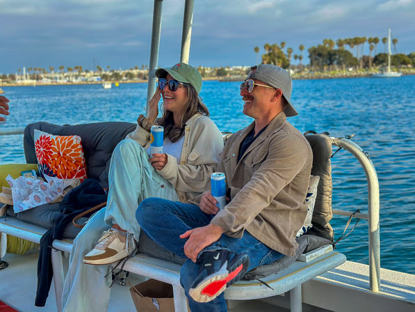 Two friends in caps and sunglasses laugh on a cushioned boat bench, holding canned drinks with a palm-lined harbor and sailboats on a sunny coastal backdrop.