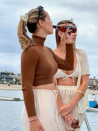 Two women on a boat near a coastal shoreline — one in a brown cropped turtleneck and white drawstring pants sipping from a bottle, the other in a white bikini and crochet cover-up holding a glass jar, overcast beach and houses in the background.