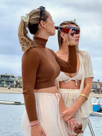 Two women on a boat near a coastal shoreline — one in a brown cropped turtleneck and white drawstring pants sipping from a bottle, the other in a white bikini and crochet cover-up holding a glass jar, overcast beach and houses in the background.