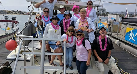 Smiling group of adults wearing pink leis and sunglasses posing with a dog on the deck of a white boat at a sunny marina dock.