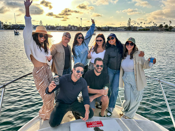 Eight friends on the bow of a boat enjoying a sunset cruise party with drinks, smiling and posing in a palm-lined coastal harbor with calm water and waterfront homes.