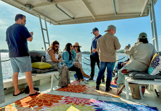 Group of friends relaxing on a sunlit covered boat deck with colorful floral rugs and cushions, chatting and sipping drinks while overlooking calm bay water and distant palm trees.