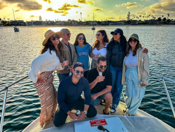 Eight friends laughing and holding drinks on a boat at sunset in a palm-lined coastal harbor with sailboats and waterfront homes.