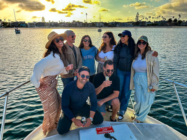 Eight friends laughing and holding drinks on a boat at sunset in a palm-lined coastal harbor with sailboats and waterfront homes.
