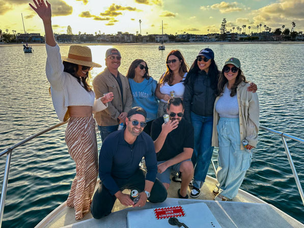 Group of eight friends enjoying a boat party on San Diego Bay at sunset, smiling and holding drinks with palm-lined shoreline and sailboats in the background.