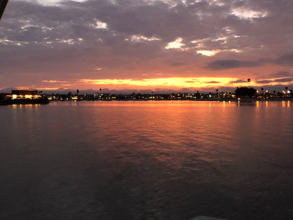 Glowing orange-pink sunset over a calm coastal bay, silhouetted palm trees and twinkling shoreline lights reflecting on rippled water