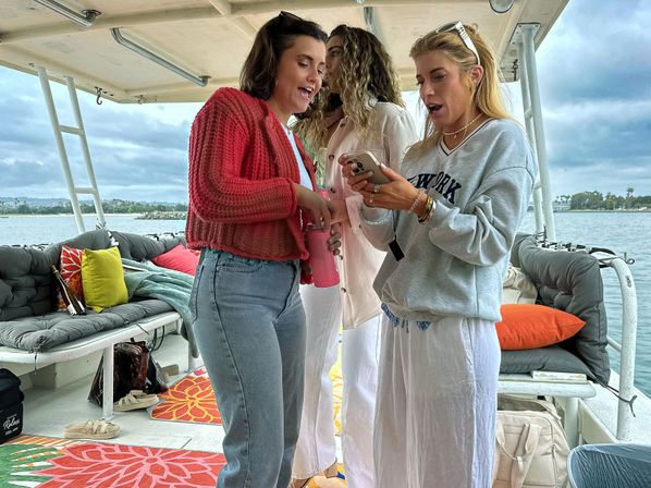 Three women on a covered boat deck in a coastal harbor, laughing and looking at a smartphone with colorful cushions and an overcast sky in the background.