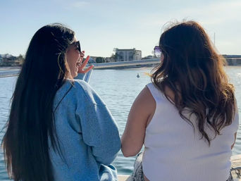 Two friends in sunglasses laughing on a sunny boat ride, sitting by calm bay waters with waterfront homes and a marina in the background.