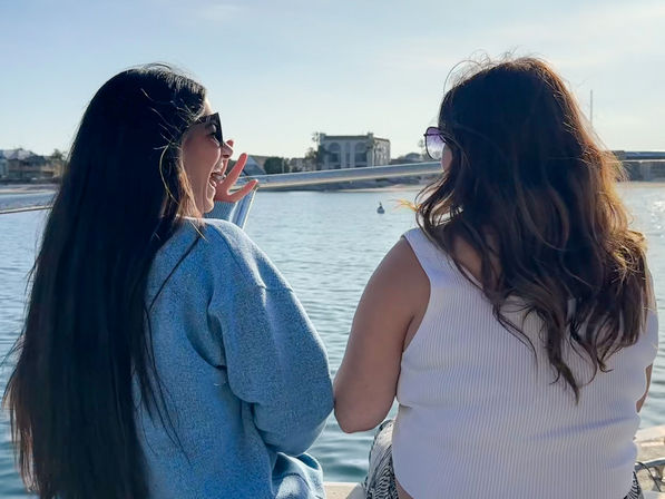 Two friends in sunglasses laughing on a sunny boat ride, sitting by calm bay waters with waterfront homes and a marina in the background.