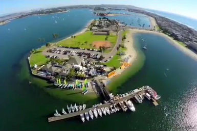 Aerial drone view of a sunny bayfront park peninsula with sandy beach, green lawns, parking lot and a marina pier lined with small sailboats and colorful dinghies.