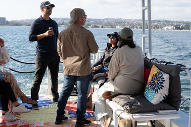 Group of people chatting on a small pleasure boat cruising a sunny bay with colorful cushions and a coastal skyline in the background