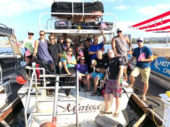Happy group on a party boat at a sunny coastal marina, waving American flags and flowers, one person holding a small dog, dock and harbor in the background