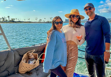 Three friends smiling on a sunny boat in a Southern California bay with palm trees on the distant shore; one woman wears a straw hat and holds a drink, a wicker tote and phone on a cushioned bench.