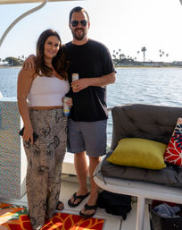 Smiling couple on a sunny boat deck holding canned drinks, standing beside cushioned seating and colorful pillows with a palm-tree-lined coastal bay and shoreline in the background.