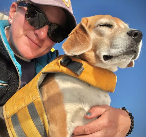 Close-up selfie of a person in sunglasses and a cap holding a relaxed beagle wearing a yellow life jacket against a clear blue sky.