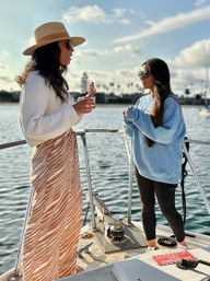 Two people chatting on the bow of a boat at a sunny marina — one in a straw hat, white sweater and patterned skirt holding a drink, the other in an oversized blue sweatshirt, black leggings and sunglasses with calm water and palm-lined harbor in the background.