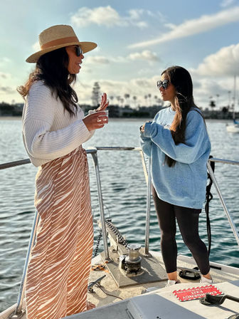 Two people chatting on the bow of a boat at a sunny marina — one in a straw hat, white sweater and patterned skirt holding a drink, the other in an oversized blue sweatshirt, black leggings and sunglasses with calm water and palm-lined harbor in the background.