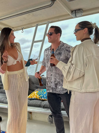 Three friends chatting and holding drinks on a boat deck with ocean view, wearing casual beachwear and sunglasses during a coastal boat outing.