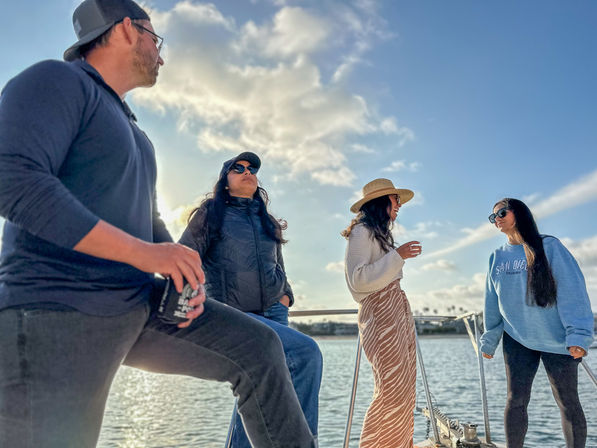 Four people chatting on a sailboat at a sunny Southern California harbor, wearing sunglasses, hats and casual layers against a blue sky.