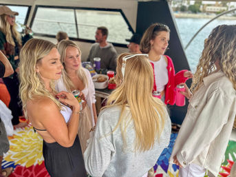 People socializing and holding drinks on a colorful-deck party boat or yacht with waterfront and shoreline visible through windows — casual coastal gathering.