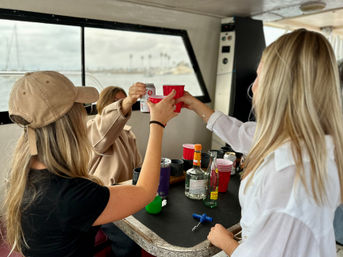 Three people toasting with red plastic cups and a canned drink around a table of liquor bottles on a boat near a palm-lined marina, coastal celebration vibe.