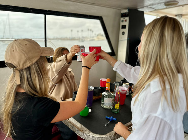 Three people toasting with red plastic cups and a canned drink around a table of liquor bottles on a boat near a palm-lined marina, coastal celebration vibe.