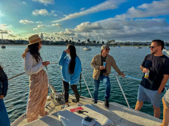 Group of friends laughing and sipping drinks on a boat deck in a sunny coastal marina with palm trees, moored sailboats and blue sky.