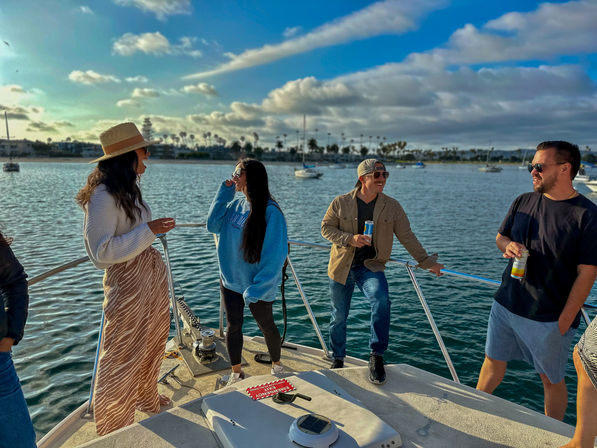 Group of friends laughing and sipping drinks on a boat deck in a sunny coastal marina with palm trees, moored sailboats and blue sky.