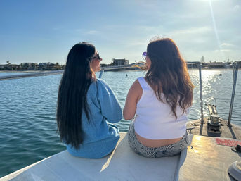 Two people sitting on a boat bow at a sunny marina, facing calm water and waterfront homes, wearing sunglasses and casual summer clothes with sparkling sunlight on the water.