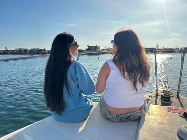 Two people sitting on a boat bow at a sunny marina, facing calm water and waterfront homes, wearing sunglasses and casual summer clothes with sparkling sunlight on the water.