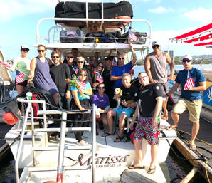 Smiling group of friends and a dog on a party boat at a sunny San Diego marina, many waving small American flags and holding flowers.
