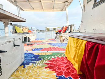 Covered boat deck with vibrant floral rugs, cushioned benches and colorful throw pillows, cozy seating overlooking a calm bay and palm-lined shoreline