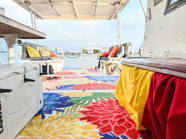 Covered boat deck with vibrant floral rugs, cushioned benches and colorful throw pillows, cozy seating overlooking a calm bay and palm-lined shoreline