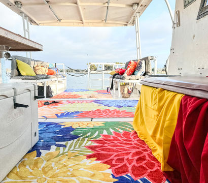 Covered boat deck with vibrant floral rugs, cushioned benches and colorful throw pillows, cozy seating overlooking a calm bay and palm-lined shoreline