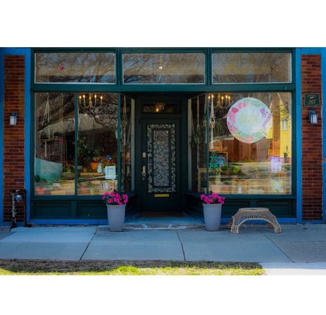 Charming main-street boutique storefront with teal trim and large display windows, chandeliers inside, two potted pink flowers flanking the door and a wicker bench on the sidewalk.