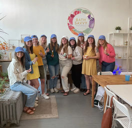 Ten women wearing matching blue caps smile in a bright boutique studio, holding natural skincare and bath products during a small-group workshop.
