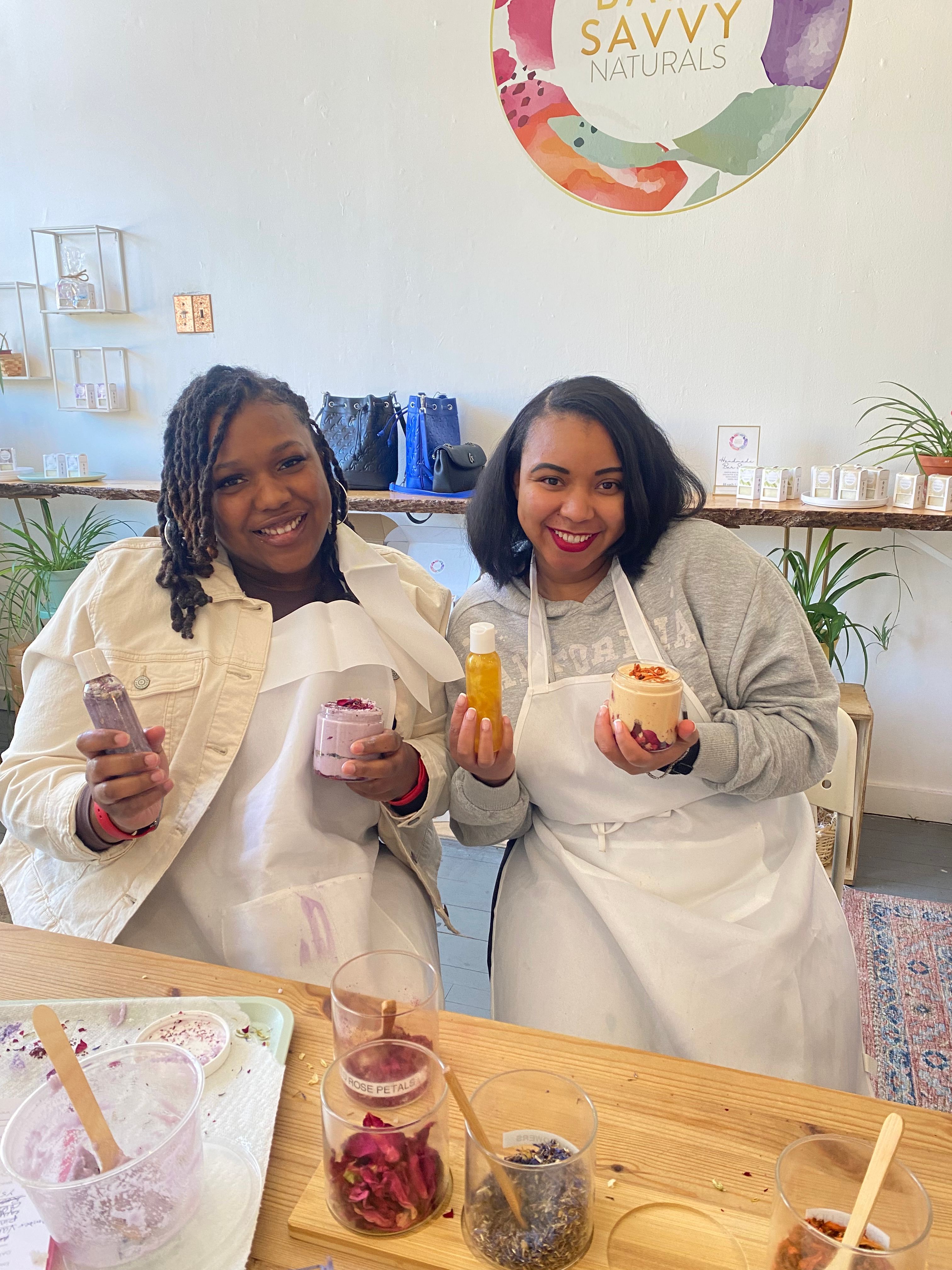 Two smiling women in aprons holding homemade natural skincare jars and bottles at a bright DIY skincare workshop, with jars of dried rose petals and botanicals on the table.